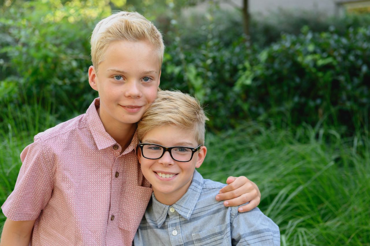 Two boys stand close together outdoors, one wearing glasses, both smiling, with an arm around each other.
