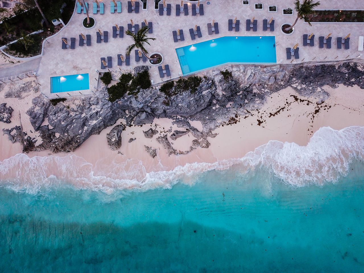 Aerial view of a beachfront resort with a swimming pool, lounge chairs, rocky cliffs, and waves washing onto the sandy shore.