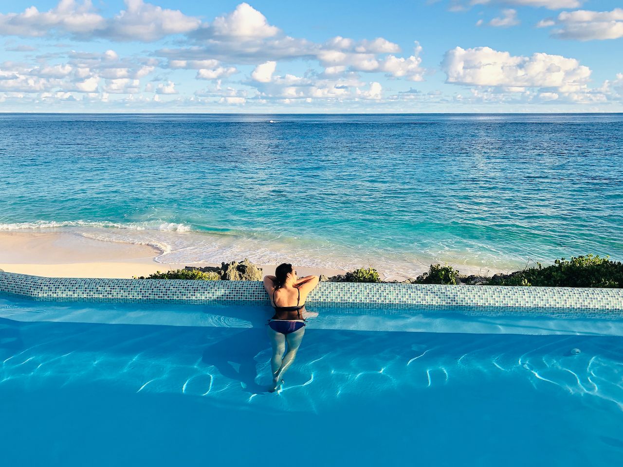 A person in a swimsuit leans on the edge of an infinity pool, looking at the ocean and beach.