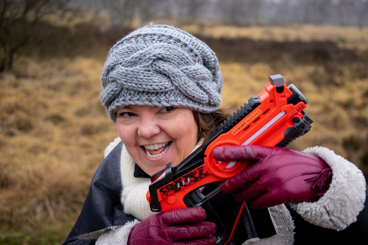 A woman wearing a gray knitted hat and gloves holds a toy gun while smiling outdoors.
