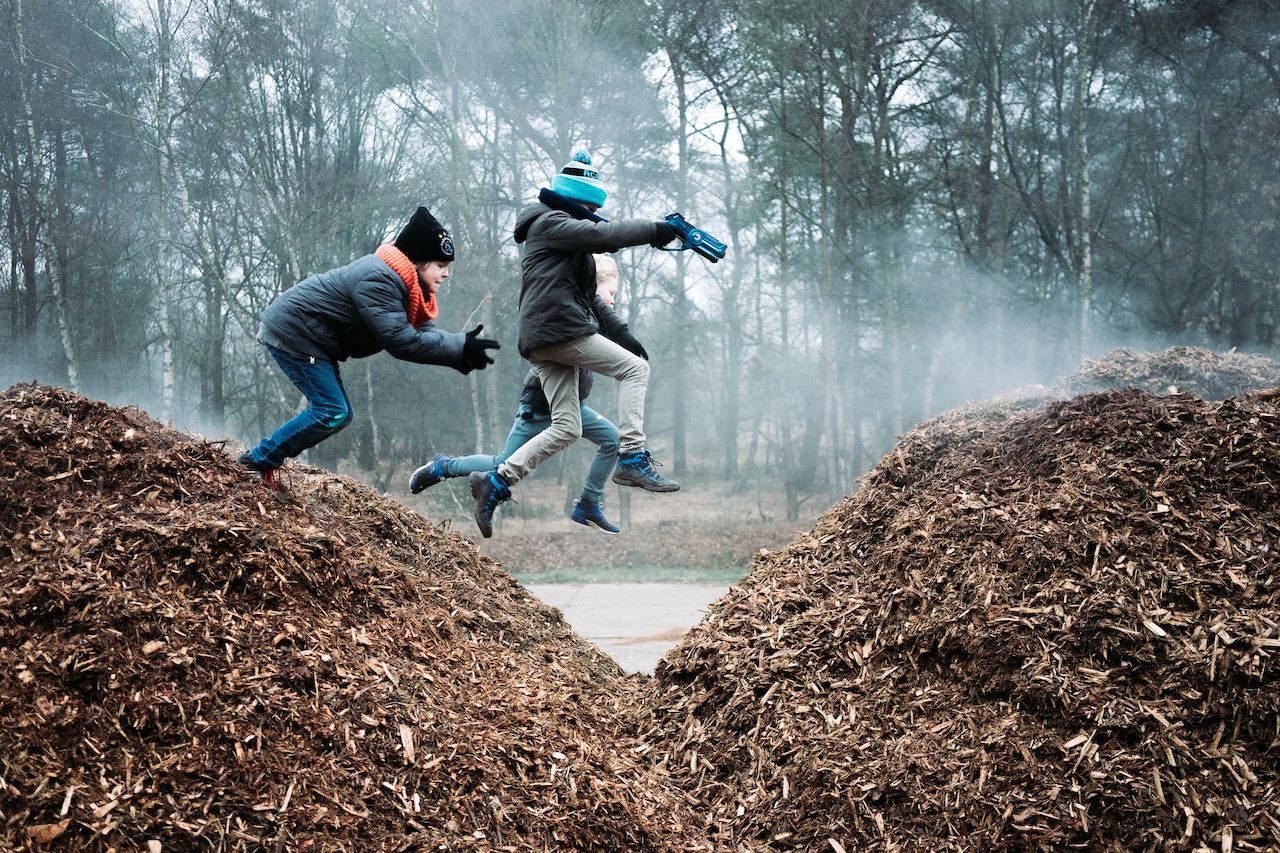 Three children in winter clothing jump between two piles of wood chips, one holding a toy blaster.