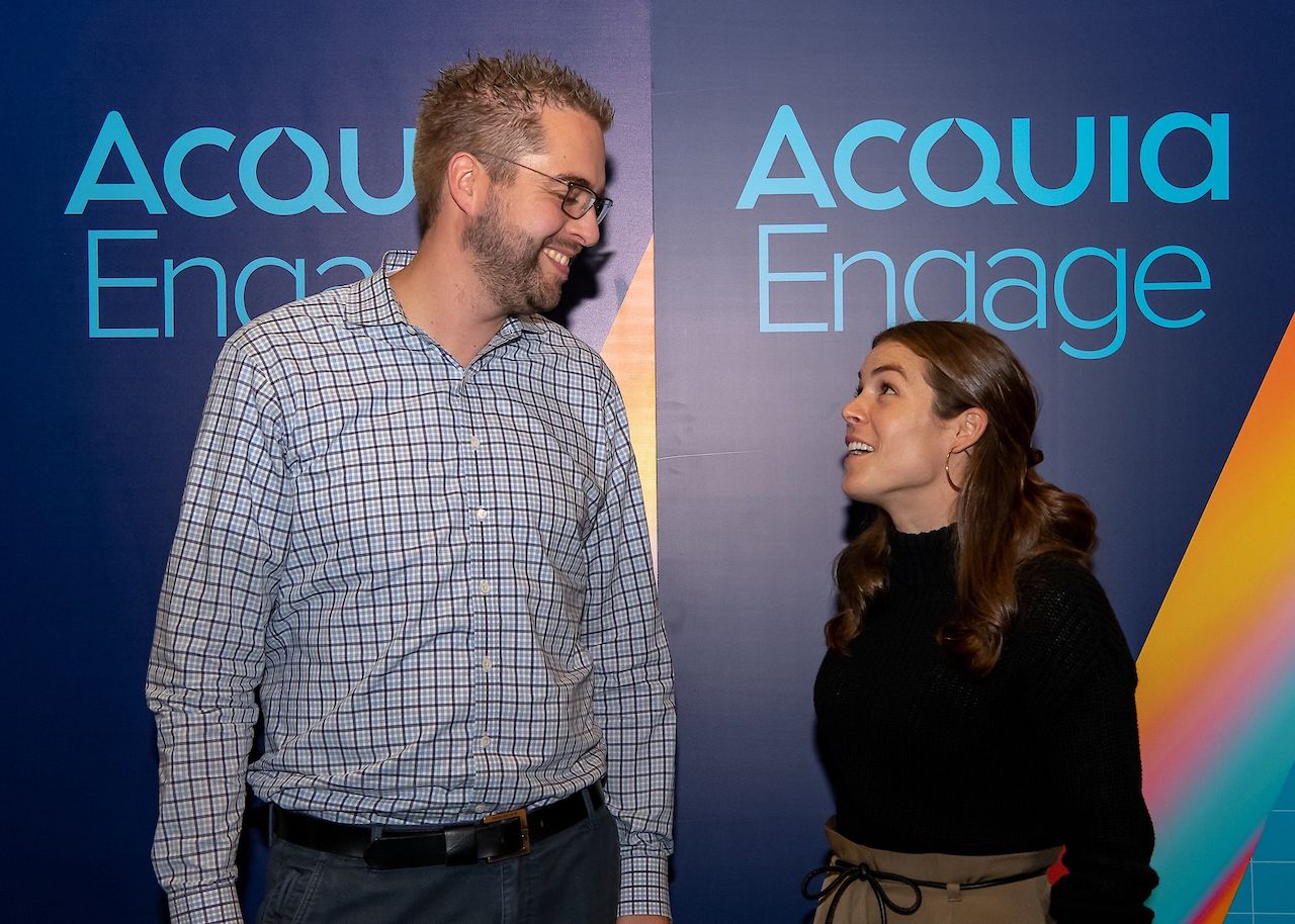 Two people stand in front of an "Acquia Engage" backdrop, smiling and talking during a meet-and-greet event.