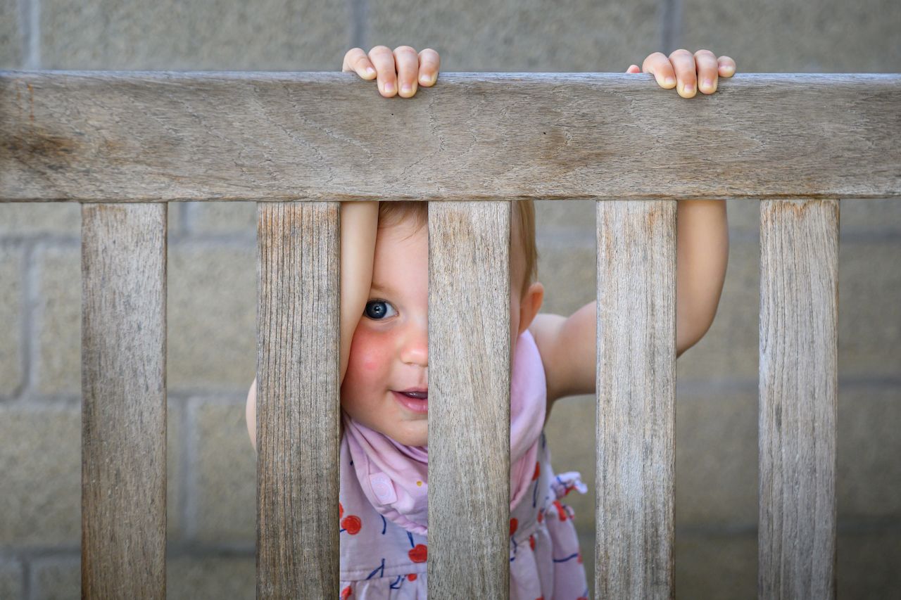 A young child peeks through the wooden slats of a bench, gripping the top rail with both hands.