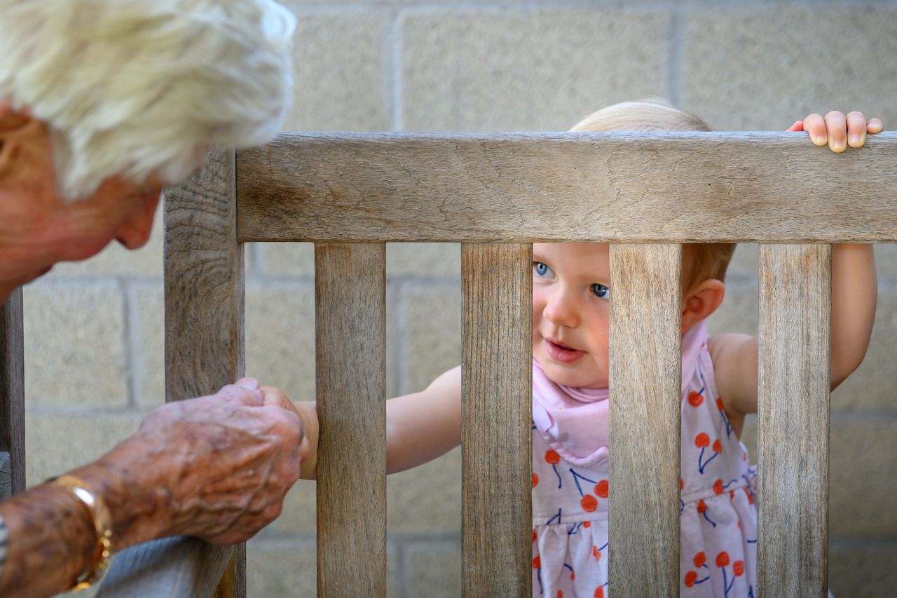 An elderly person holds a young child's hand through a wooden bench as the child looks up curiously.