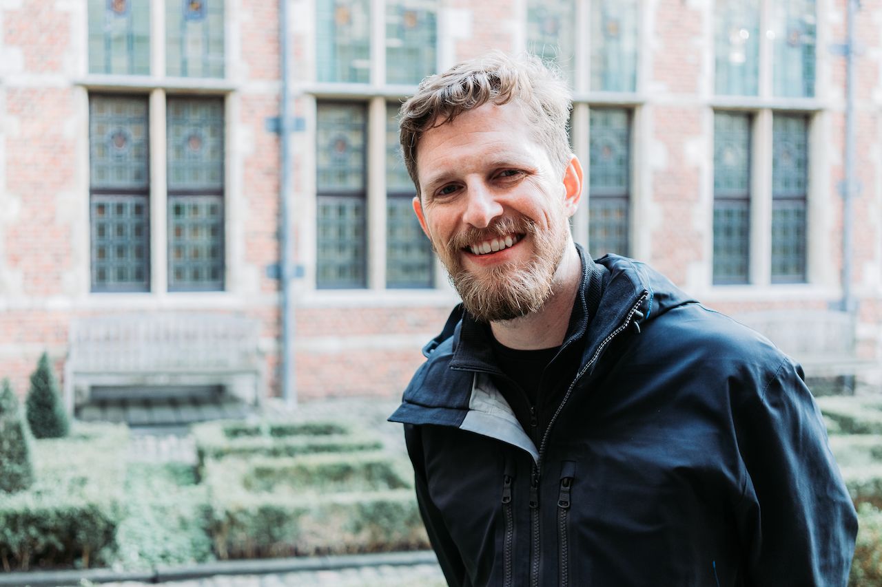 A man with a beard and a black jacket smiles in front of a historic brick building with large windows.