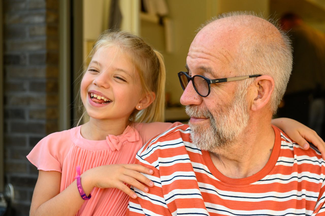 A young girl laughs while resting her arm on an older man's shoulder as he looks at her and smiles.