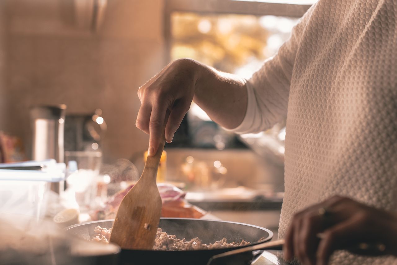 A person wearing a white sweater stirs food in a steaming pan with a wooden spatula in a kitchen.