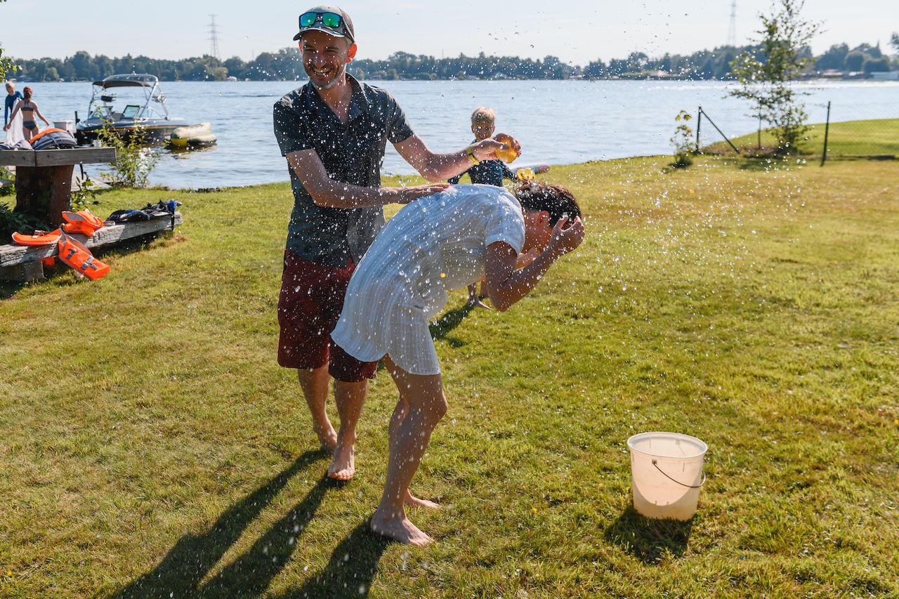 A man splashes water on another person bending forward, while a child in the background holds a water balloon.