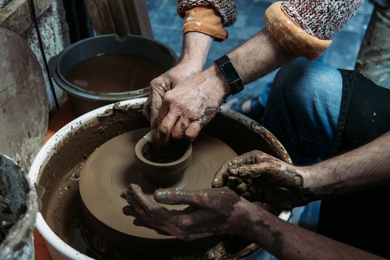 Two people shape a clay pot on a spinning pottery wheel, their hands covered in wet clay.