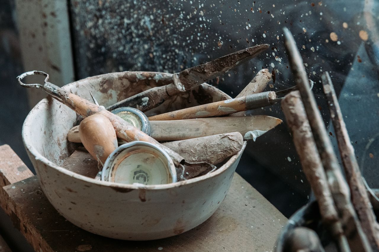 A ceramic bowl filled with well-used pottery tools, covered in dried clay, sits on a workbench.