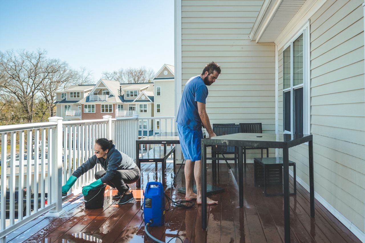 Two people cleaning a wooden deck–one scrubbing the railing, the other using a pressure washer on the floor.