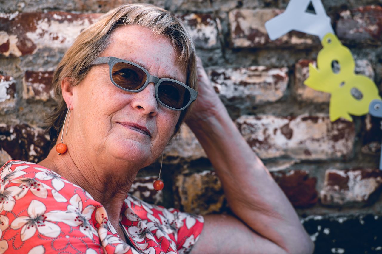 A woman wearing sunglasses and a floral shirt leans against a brick wall, resting her hand on her head.