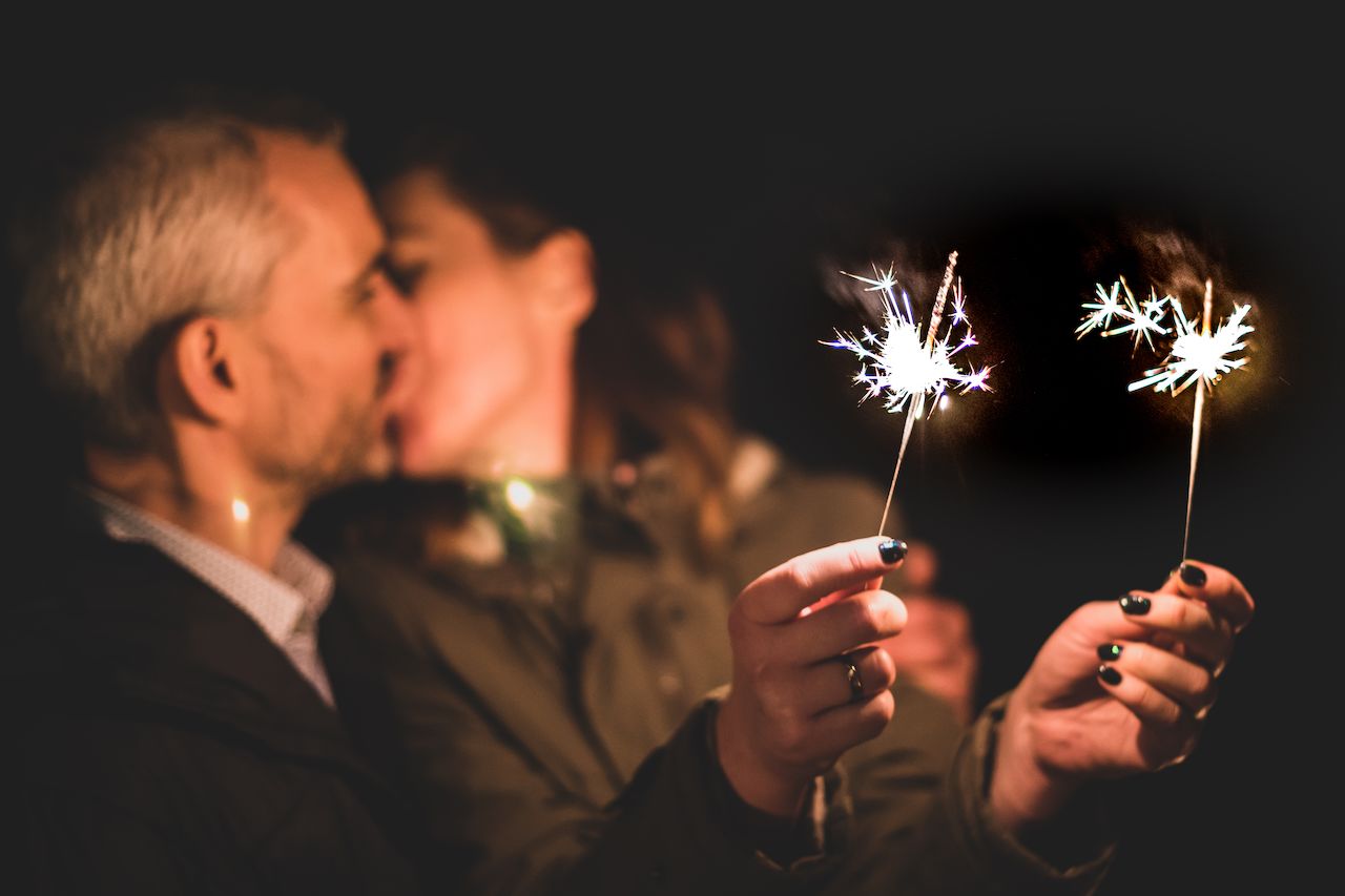 A couple shares a kiss while holding sparklers, celebrating New Year's Eve in a dark setting.
