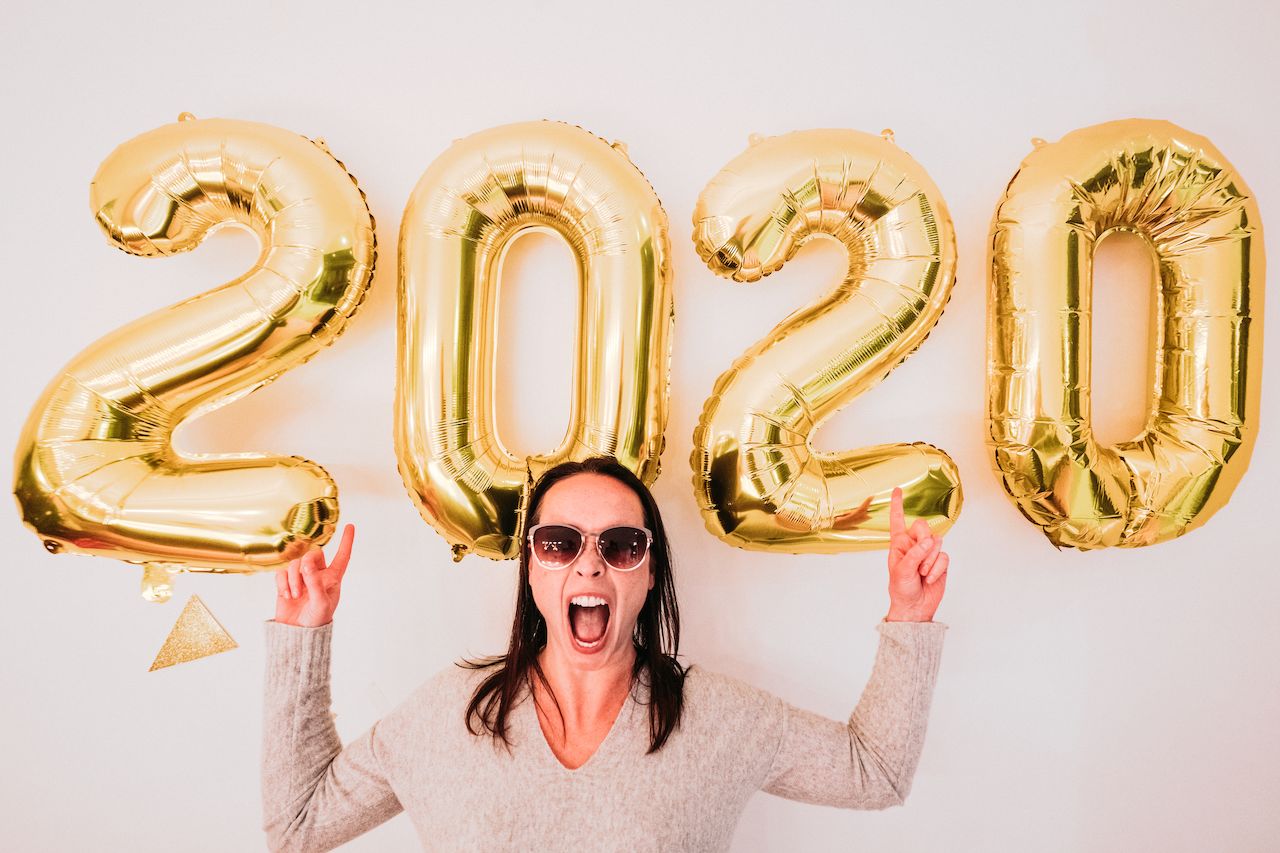 A woman wearing sunglasses smiles and raises her hands in front of large gold balloons displaying "2020.