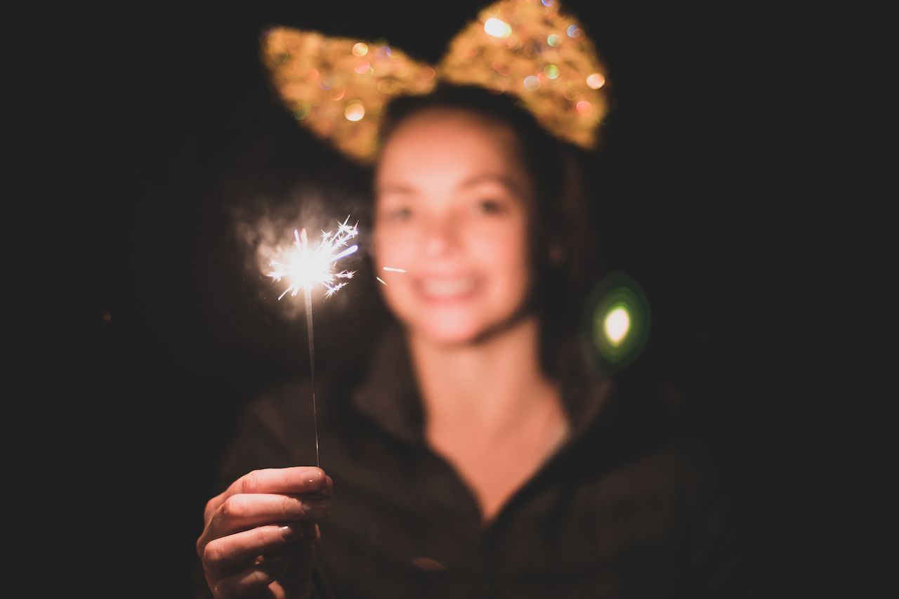 A person holds a lit sparkler while smiling, wearing a headband with glittery decorations against a dark background.