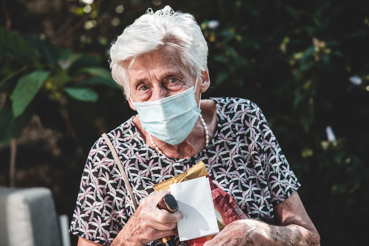 Elderly woman wearing a mask holds a small package and a card while standing outdoors.