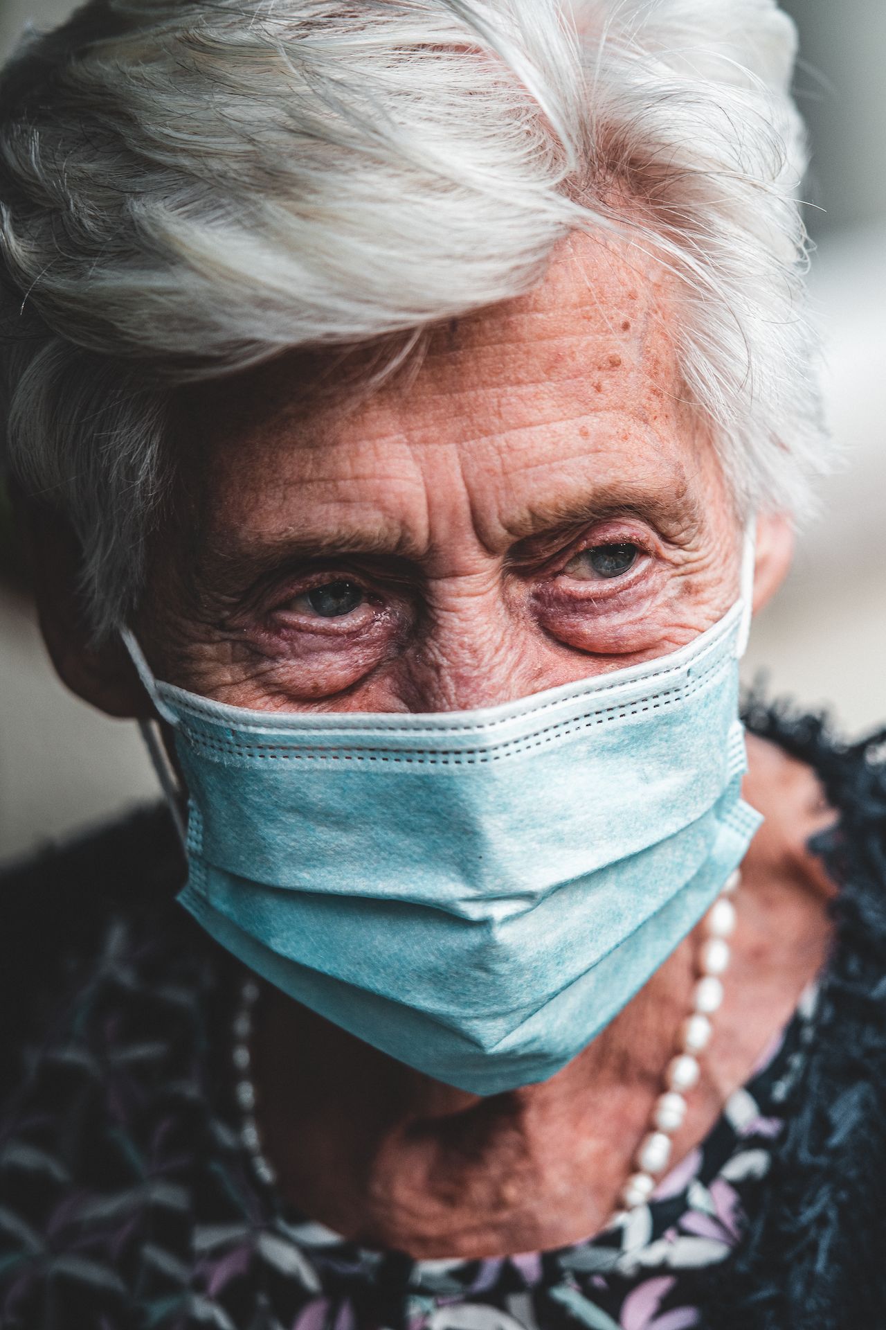 Elderly woman with white hair wearing a blue face mask and a pearl necklace, looking to the side.