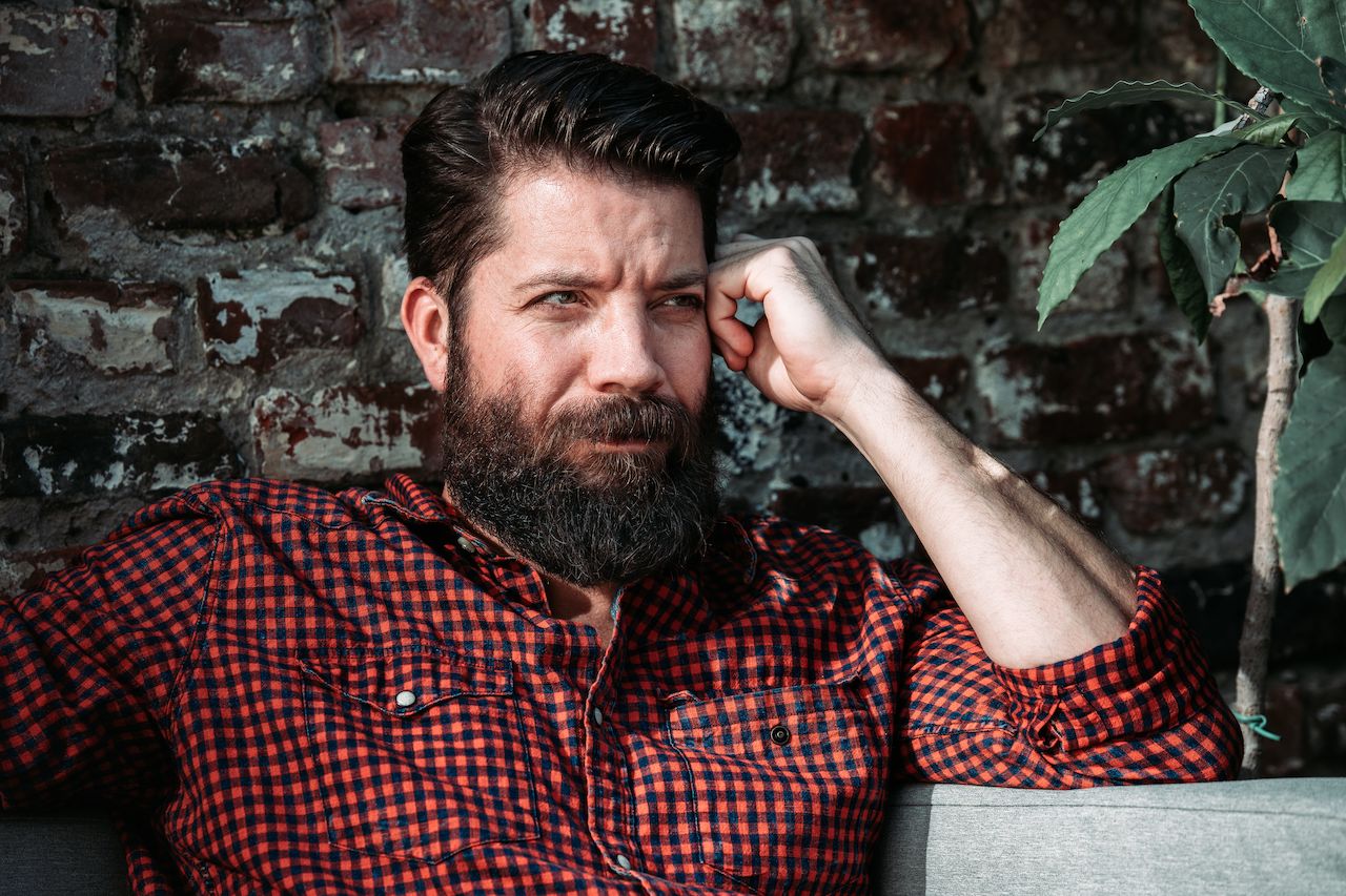 A bearded man in a red checkered shirt sits against a brick wall, resting his head on his hand.