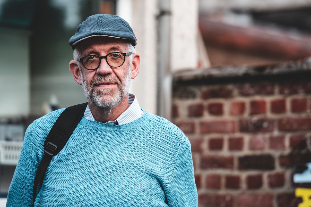 An older man wearing glasses, a cap, and a blue sweater stands outside with a bag over his shoulder.