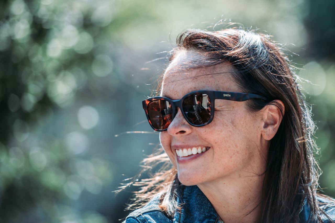 A woman wearing sunglasses and a denim jacket smiles while looking off to the side.