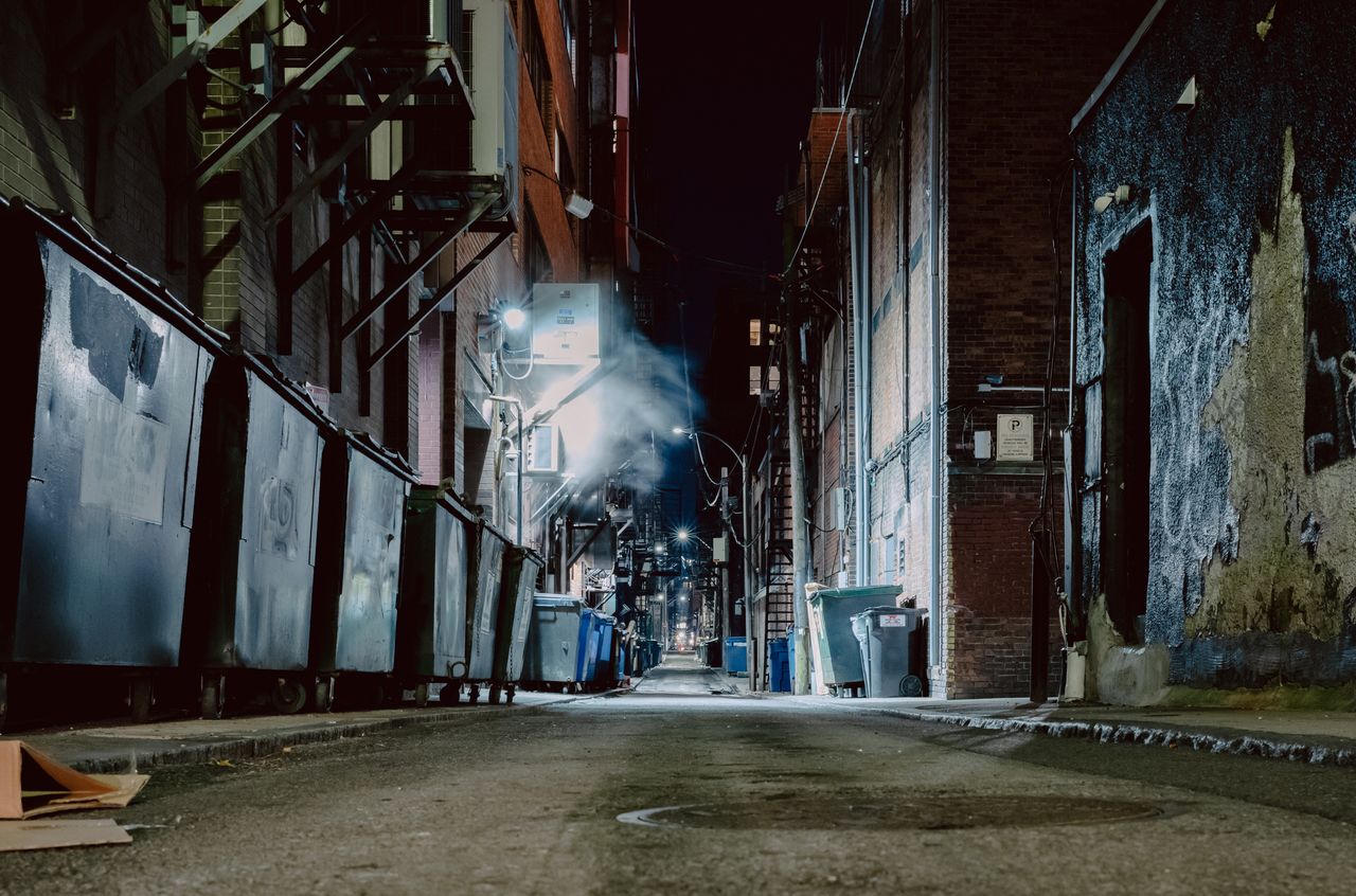 A dimly lit alley at night with dumpsters, brick buildings, and steam rising from a vent.