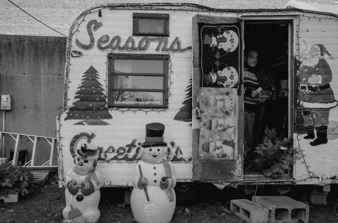A man stands inside a decorated Christmas trailer with snowman figures and festive lights outside.