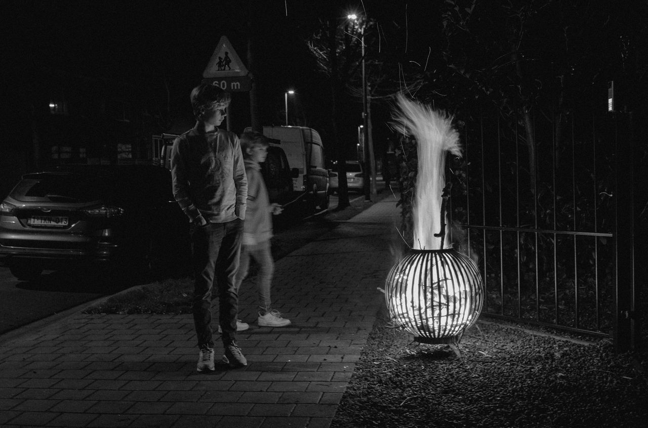 Two people stand near a fire pit with tall flames on a sidewalk at night.