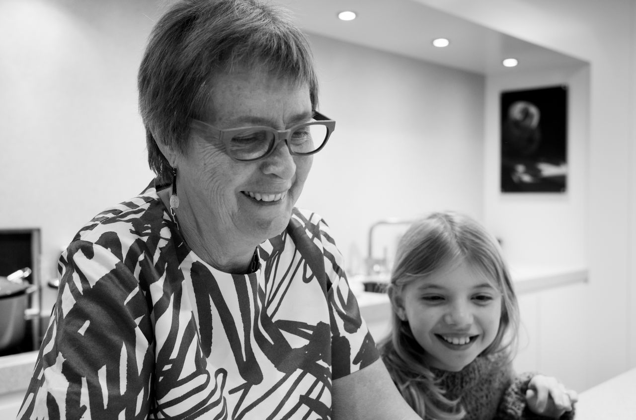 An older woman and a young girl smile together in a kitchen, looking at something in front of them.