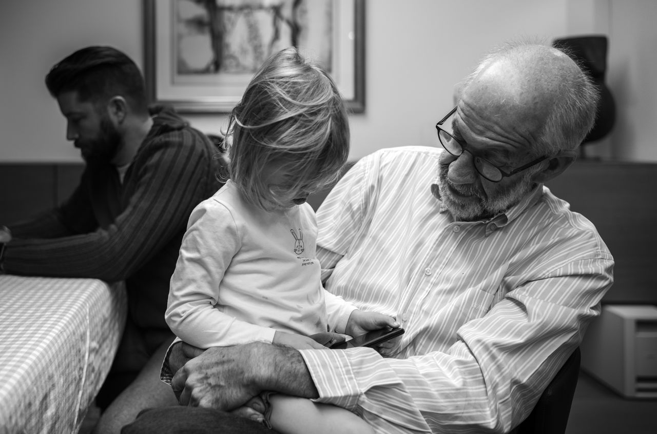 An elderly man holds a young girl on his lap as they look at a smartphone together.