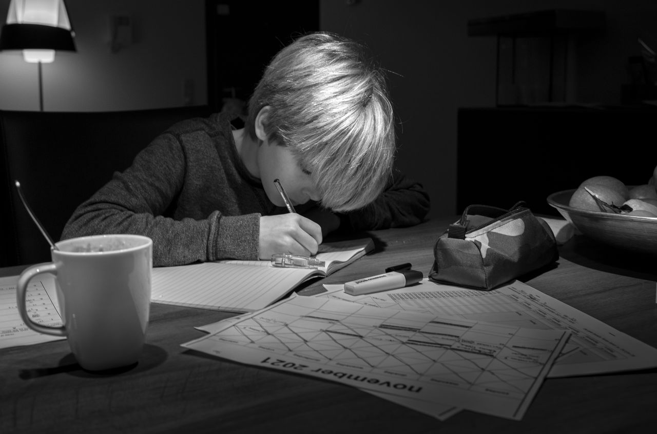 A young boy sits at a table, writing in a notebook with a pen, focused on his homework.