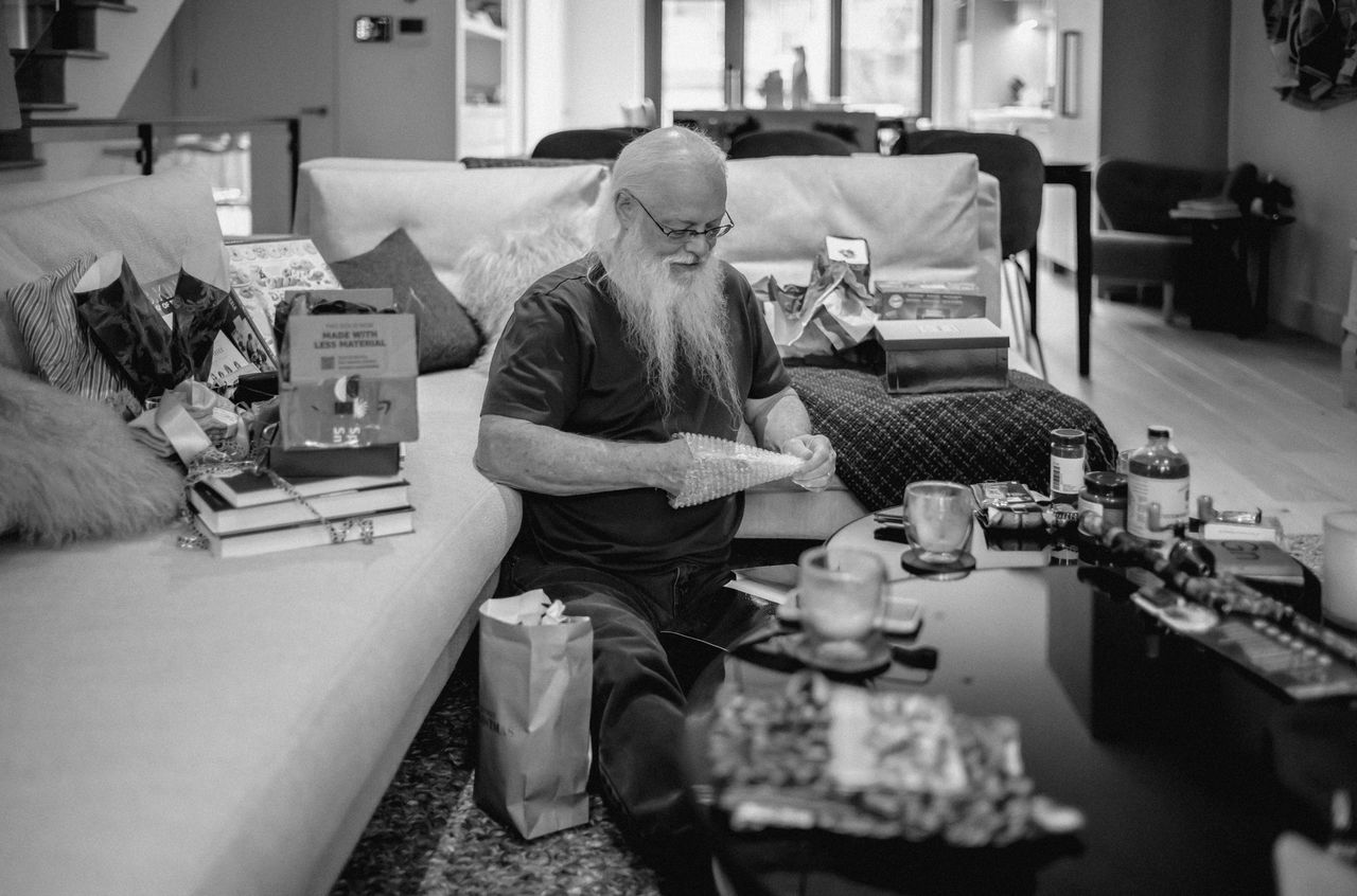 An elderly man with a long beard sits on the floor, unwrapping a gift surrounded by presents and packaging.