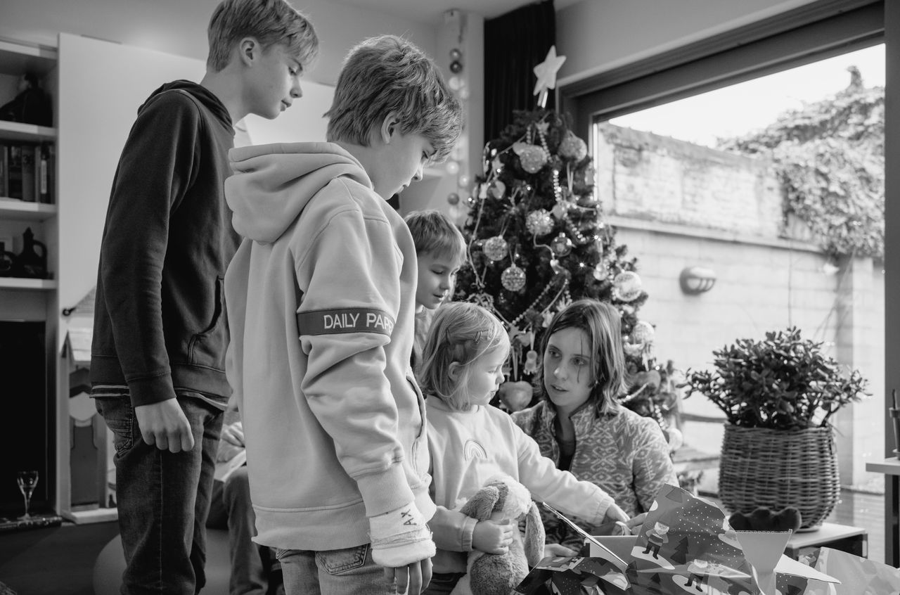 A group of children and an adult gather around a young girl as she unwraps a gift near a Christmas tree.