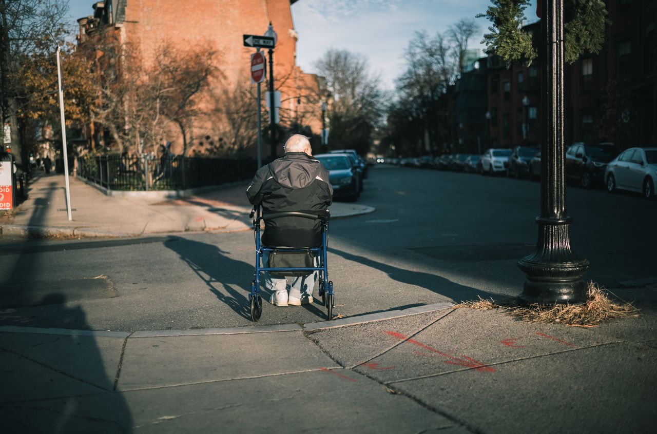 An older adult in a wheelchair moves along a sidewalk toward a street intersection in an urban setting.