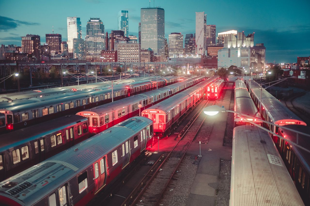 Multiple red subway trains are parked in a train yard near Boston's South Station, with the city skyline behind them.