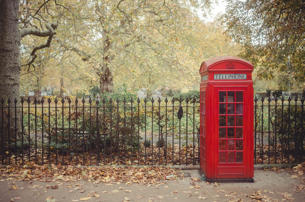 A classic red London phone booth stands in front of a fence that surrounds a local park.