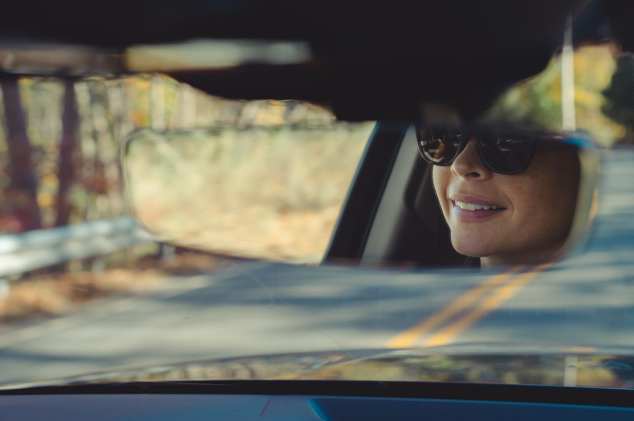 A smiling face in the rearview mirror of a car, and a road with yellow lines stretches out through the windshield.
