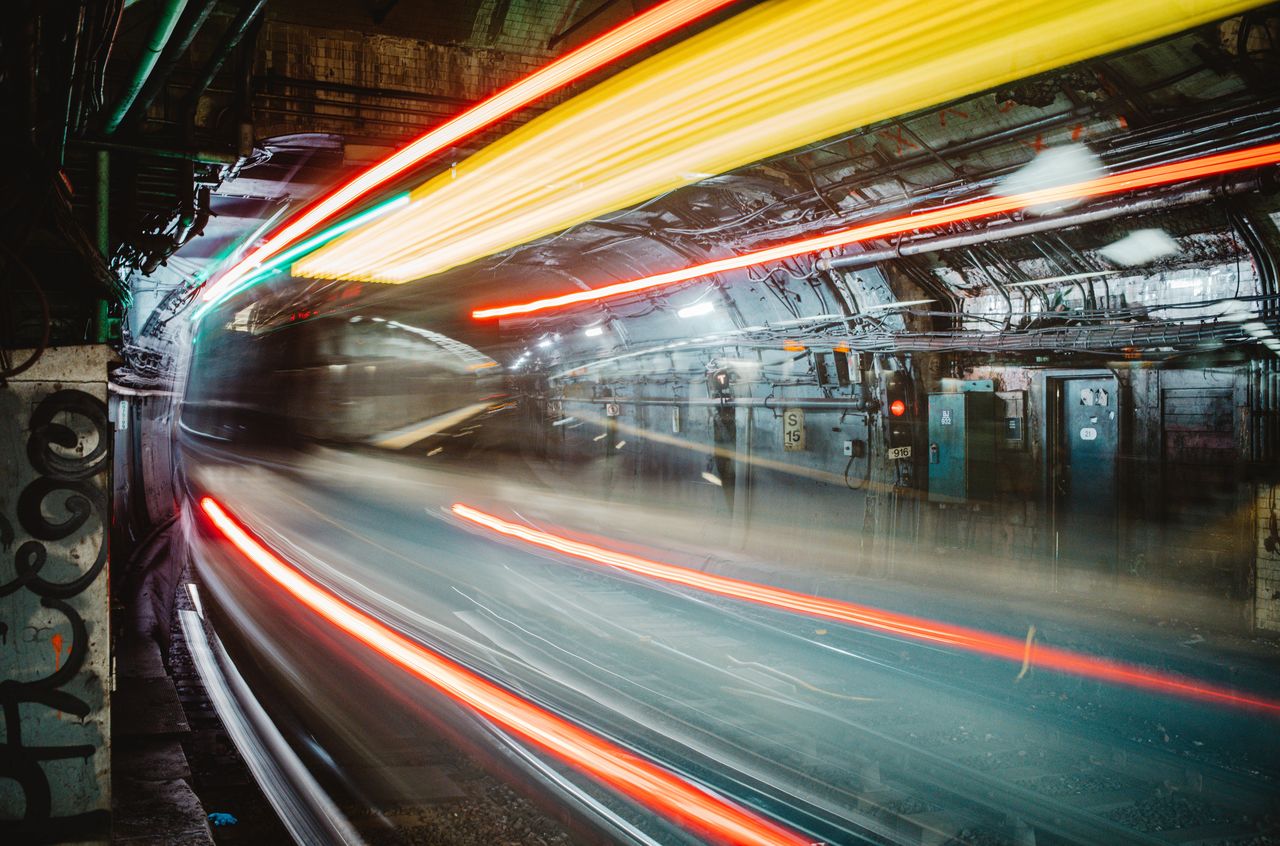 A train moves quickly through a dimly lit underground tunnel, leaving streaks of light in its path.