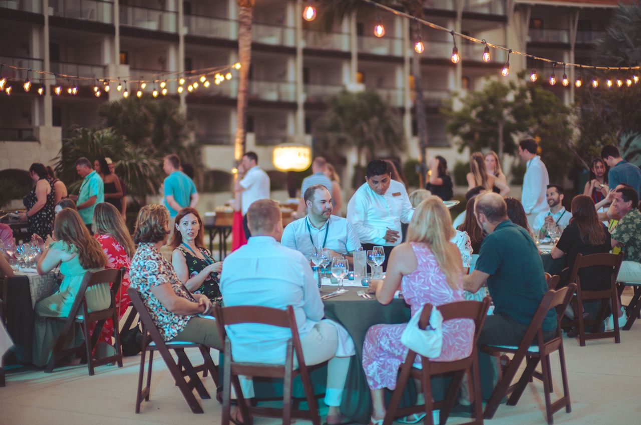 Acquia employees enjoying an outdoor dinner, seated at round tables below lights strung between palm trees.