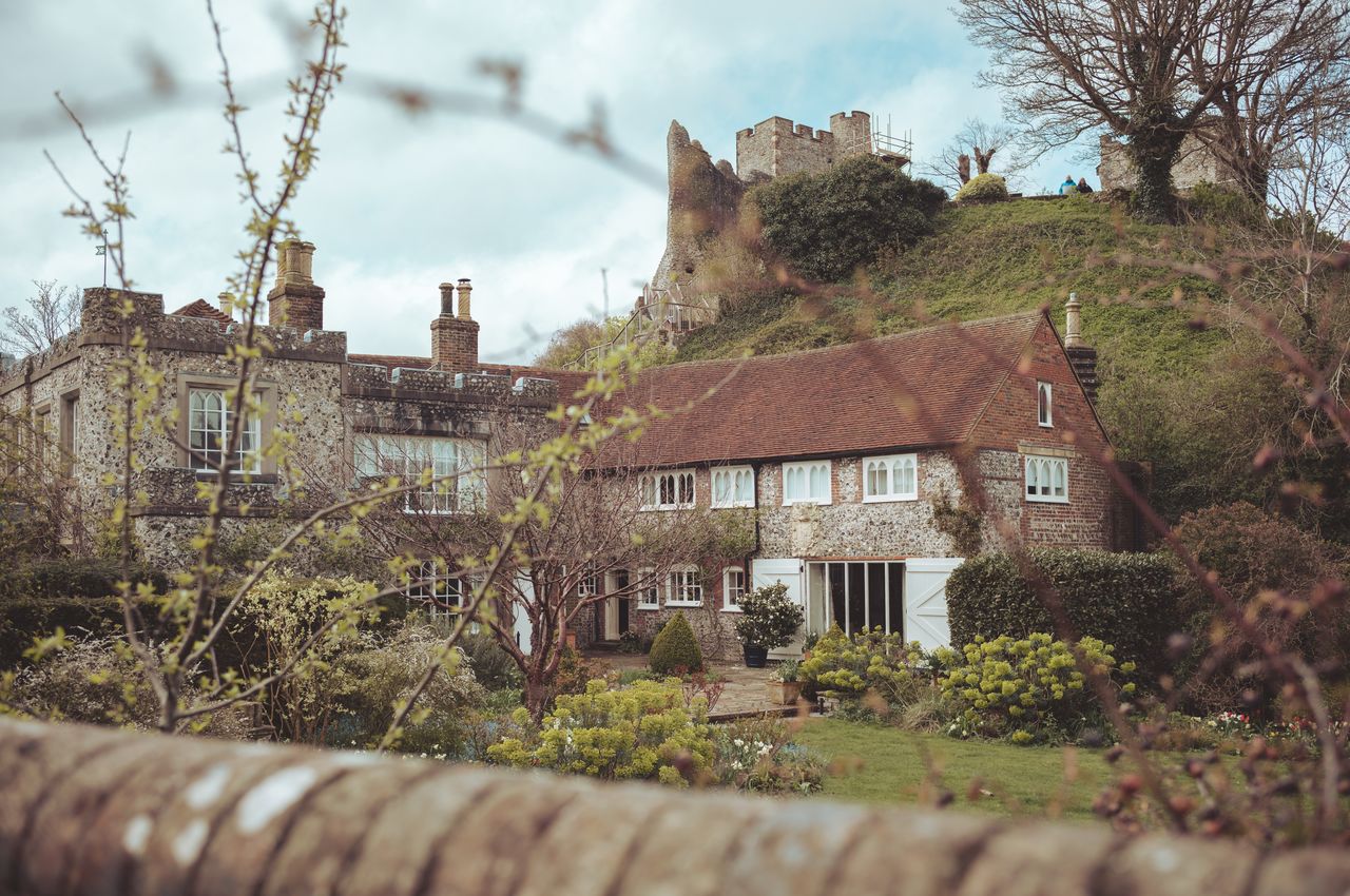 A quaint old cottage nestled at the base of a hill, surrounded by trees and flowers.