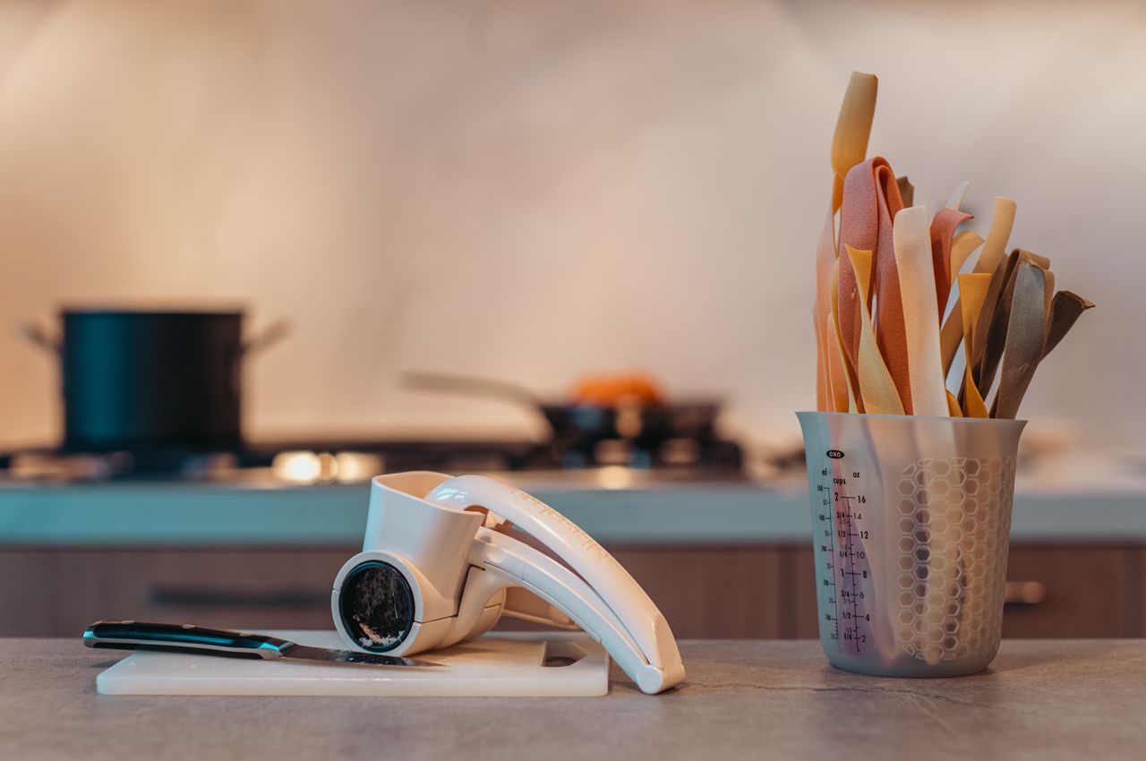 In the foreground, a cheese grater and a measuring cup holding dried pasta. In the background, a pot with hot water and a pan with bolognese sauce.