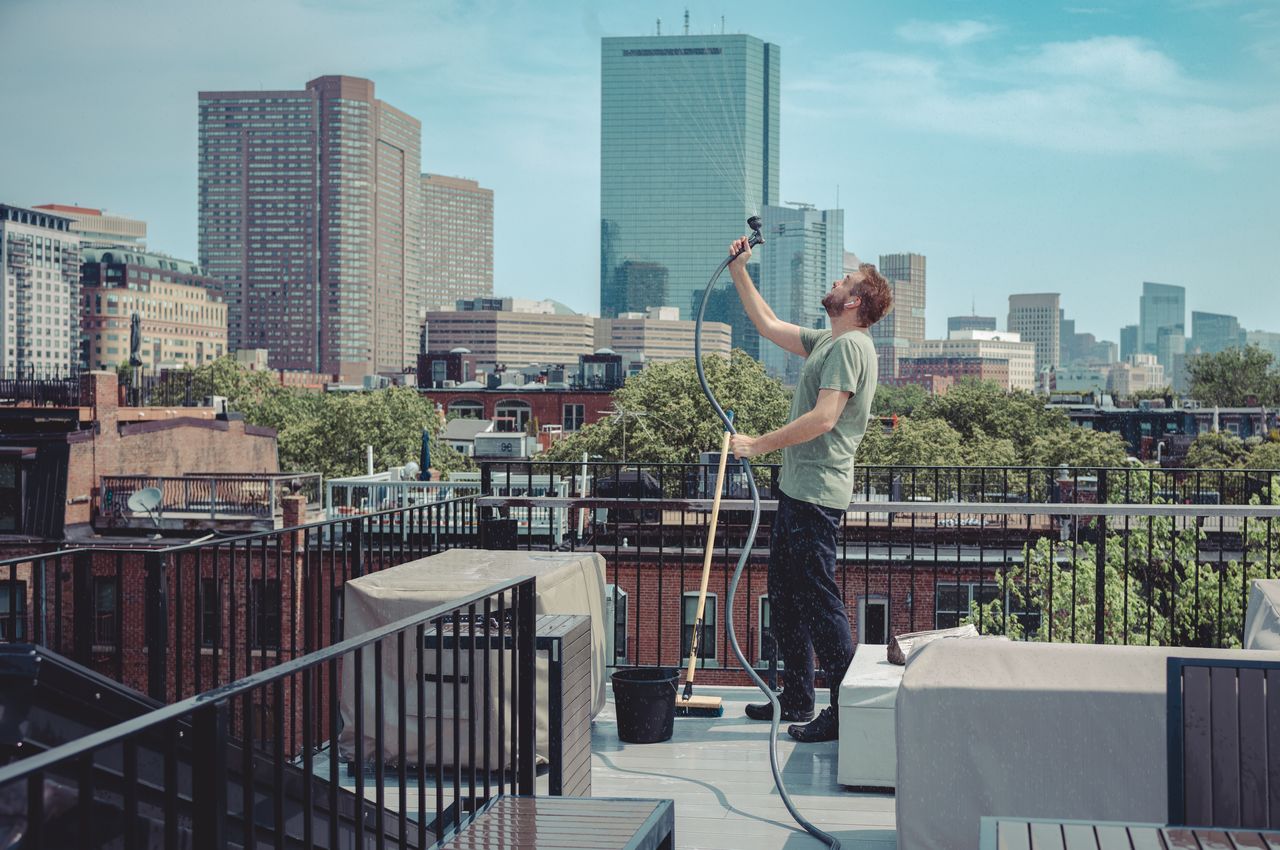 Dries is cleaning a roofdeck with the Boston skyline in the background, but then gets distracted and starts playfully spraying water into the air instead of cleaning.