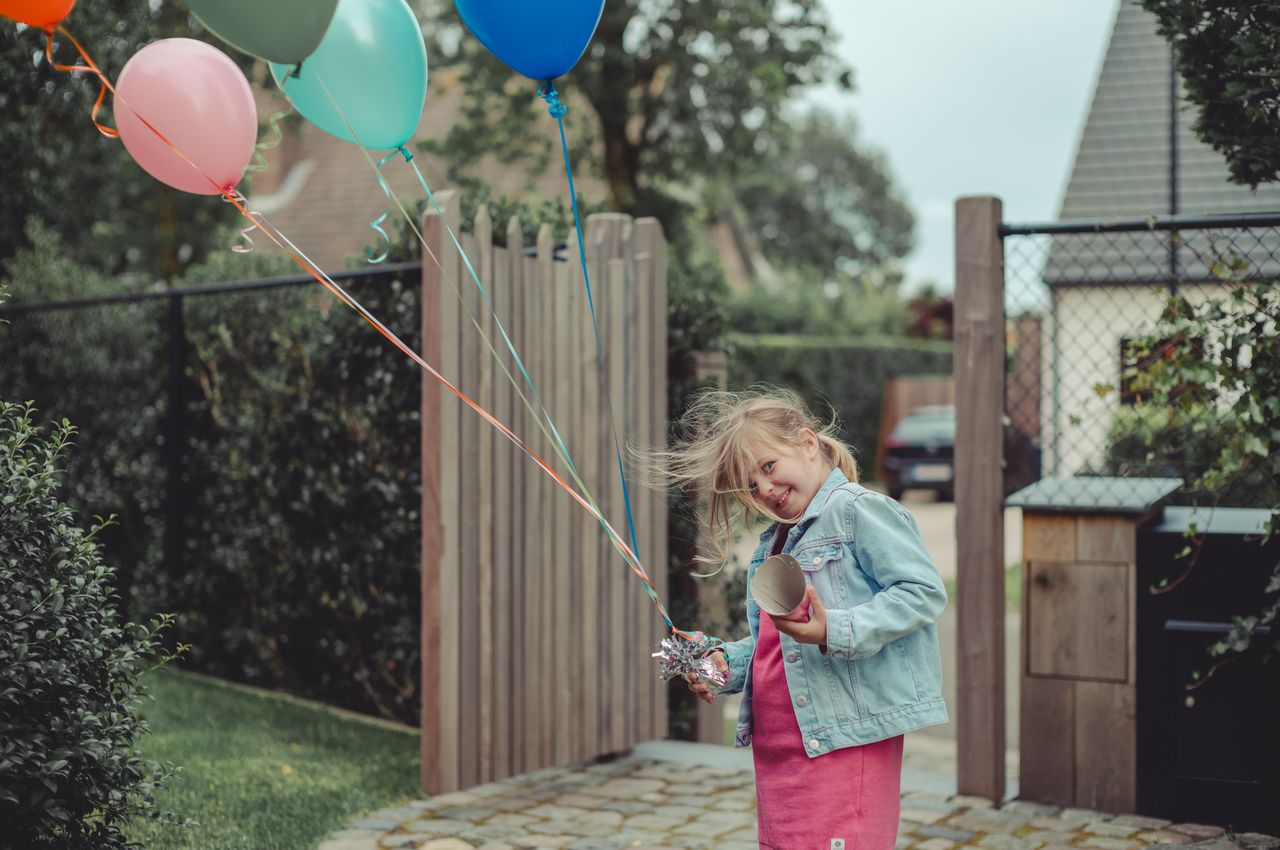 A young girl in a pink dress and denim jacket holding colorful balloons.