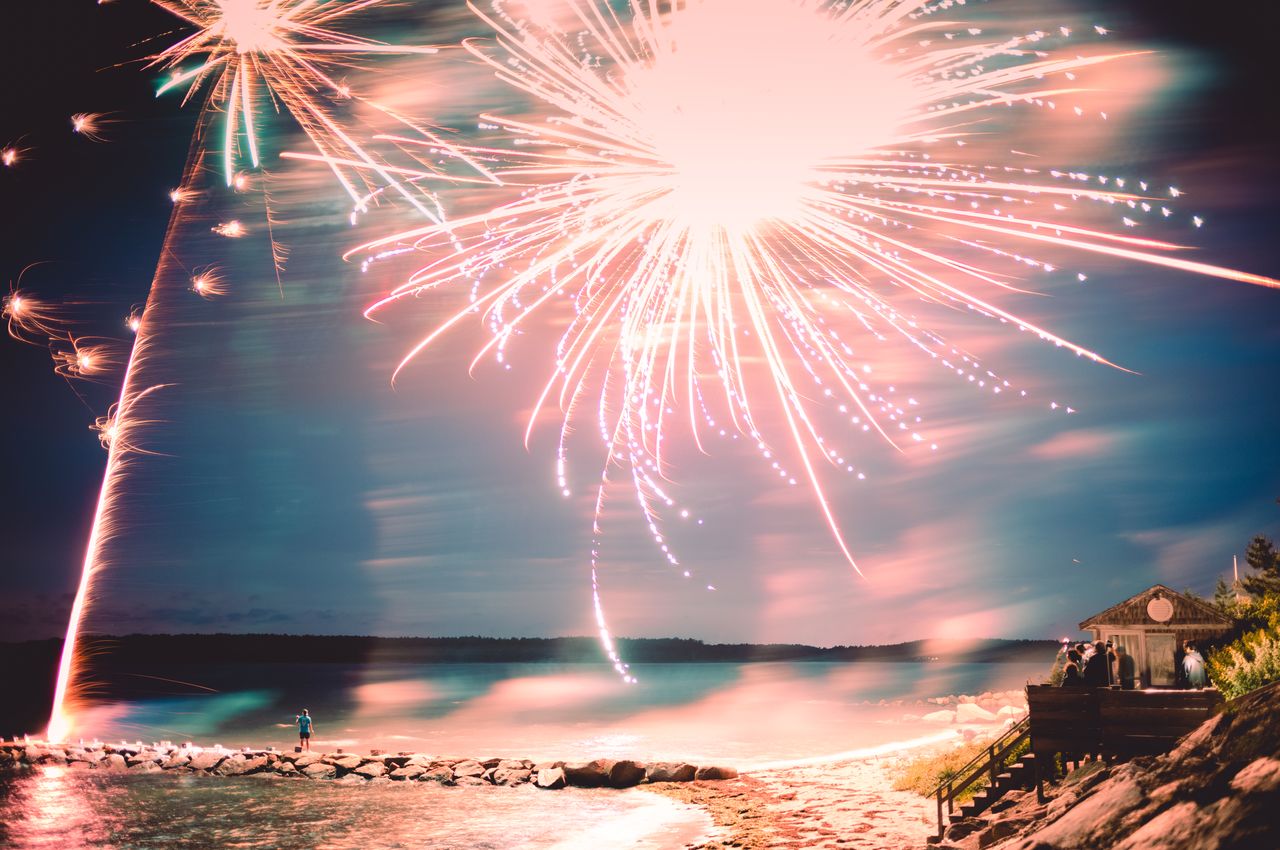 Fireworks explode over the ocean, ignited by a person standing on a jetty that extends into the water.