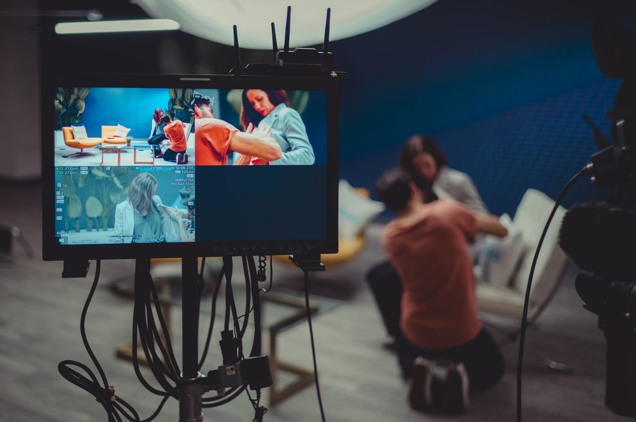 A camera monitor show a behind-the-scenes view of a woman being set up with a microphone.