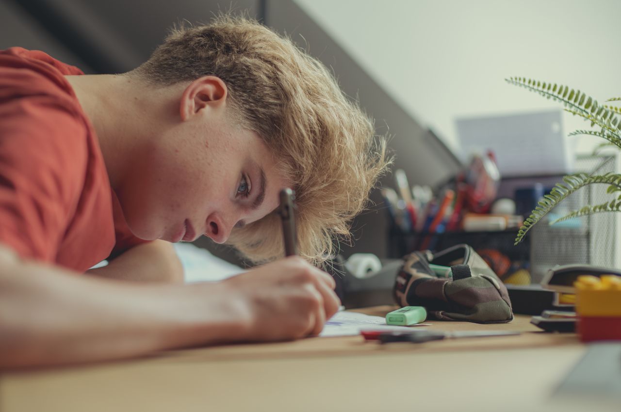 A young man sitting at a desk, head bent over study materials, focused on writing.