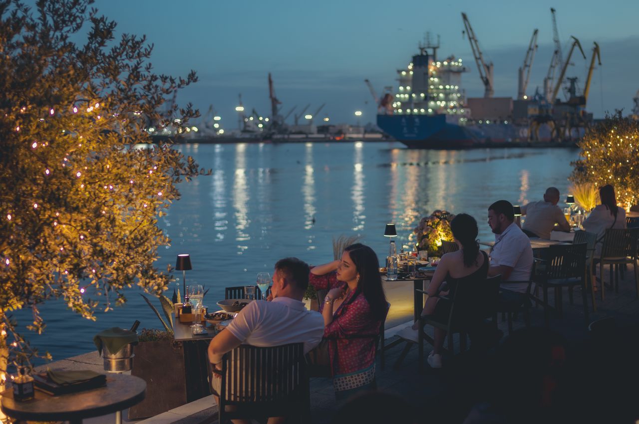 People dining outdoors with a view of the illuminated harbor and ships.