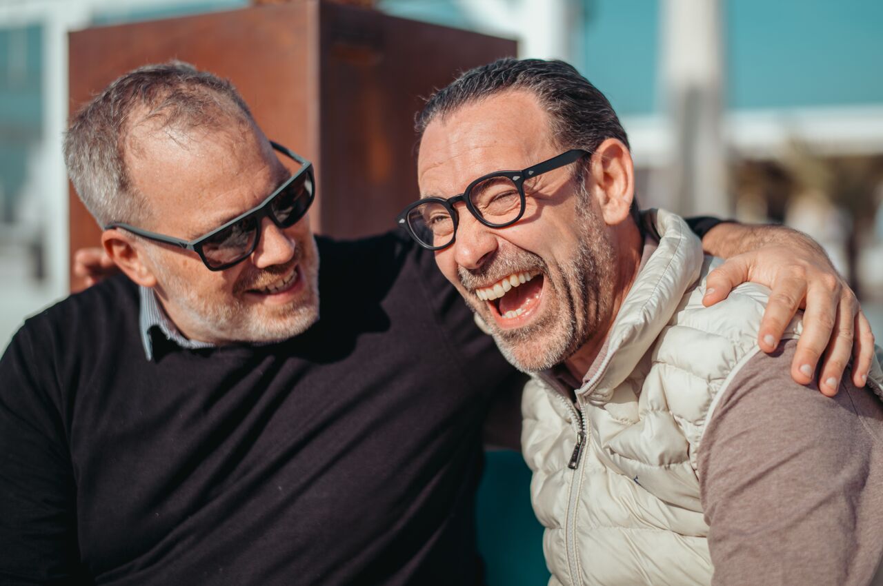 Two men laughing in the sun with arms around each other, both wearing sunglasses.