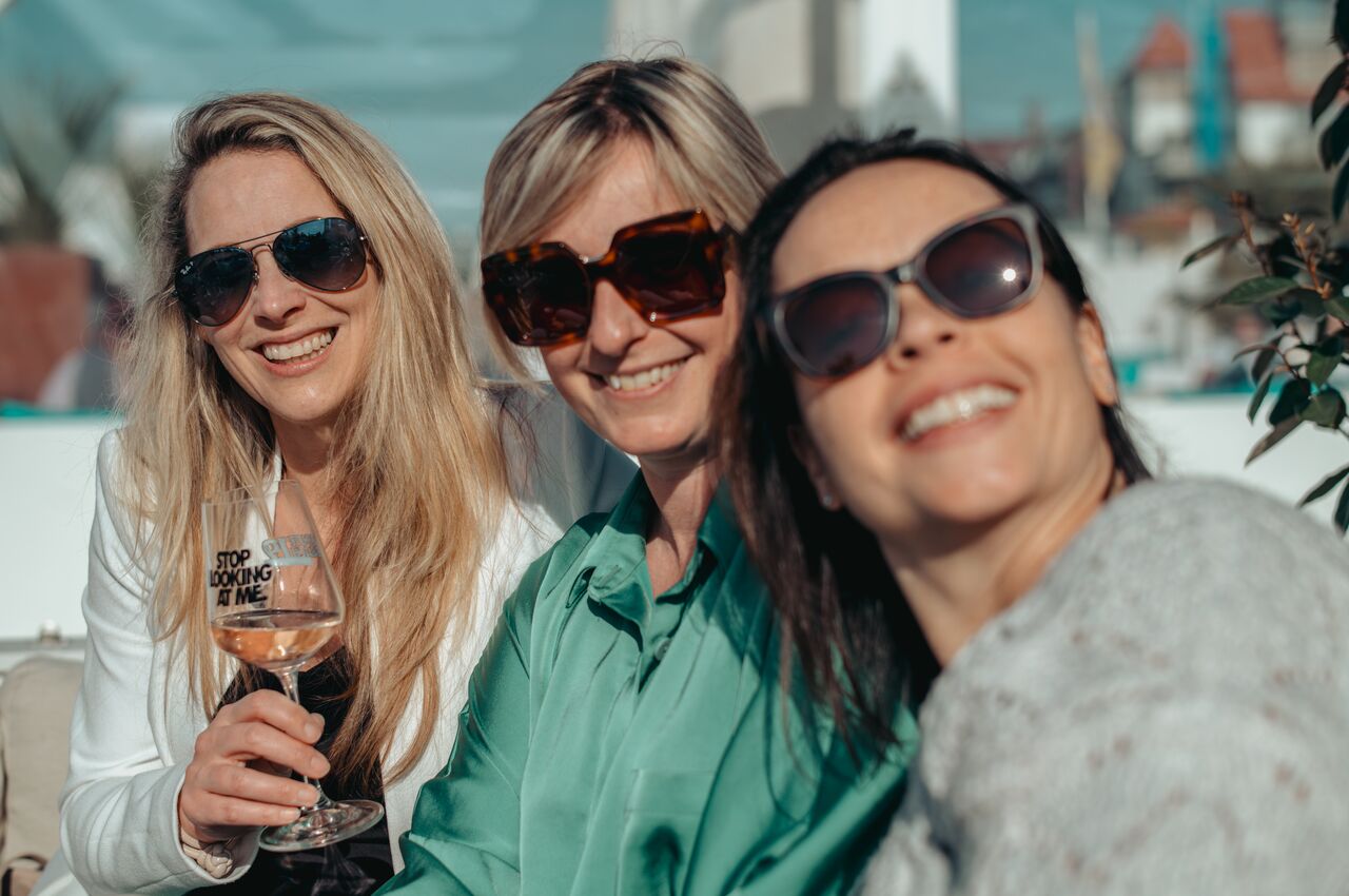Three women smiling in the sun, wearing sunglasses, with one holding a wine glass