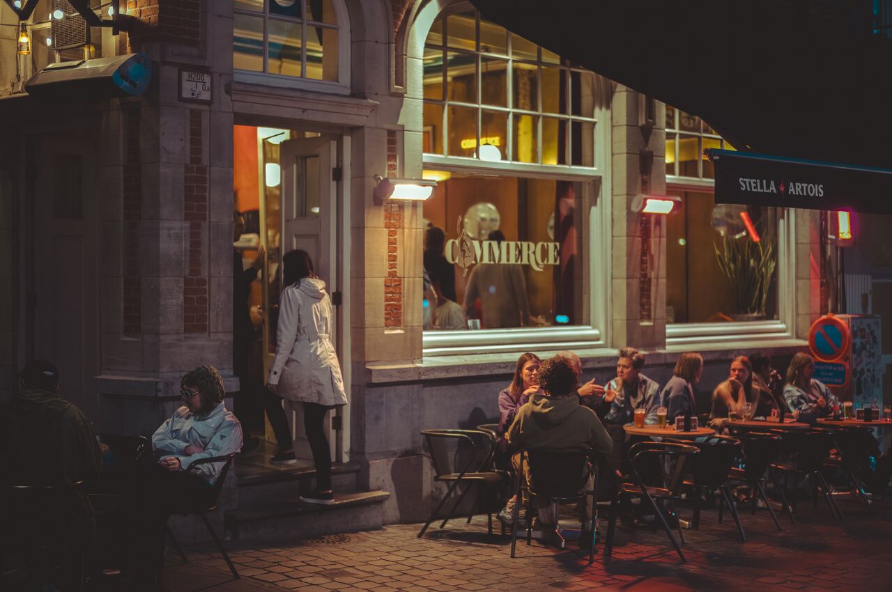 A nighttime view of a bar with "Commerce" printed on its windows, people drinking at outdoor tables, and a person entering the doorway.