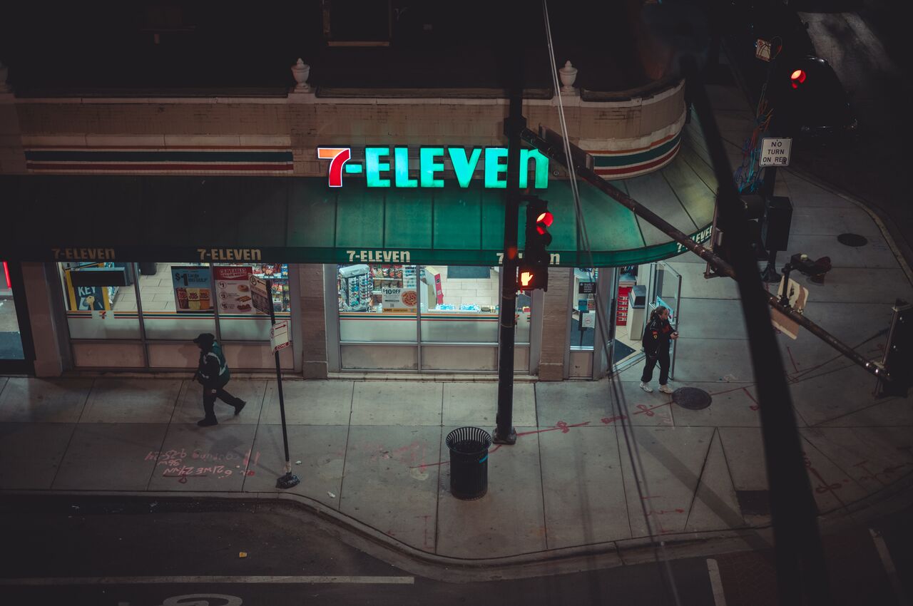 Nighttime view of a 7-Eleven store on a street corner with people walking by.
